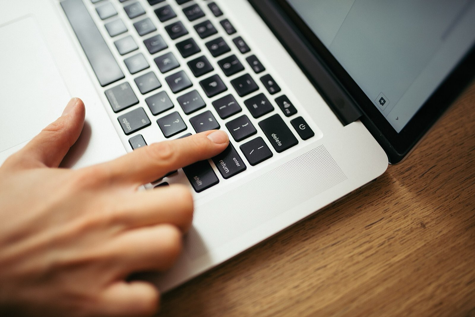 A hand is pressing keys on a laptop keyboard. The sleek device is positioned on a wooden desk, suggesting a focused work or study environment.