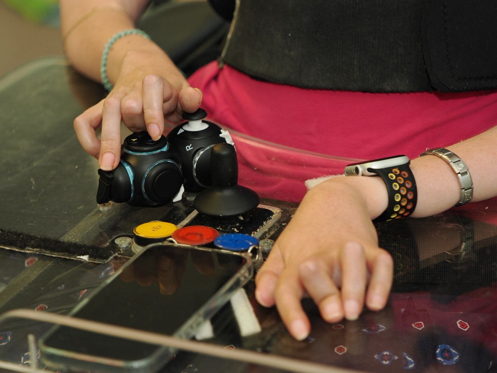 A person in a wheelchair operates a modular Proteus controller with joystick and button modules on a table; a smartphone and large colored buttons are placed nearby.